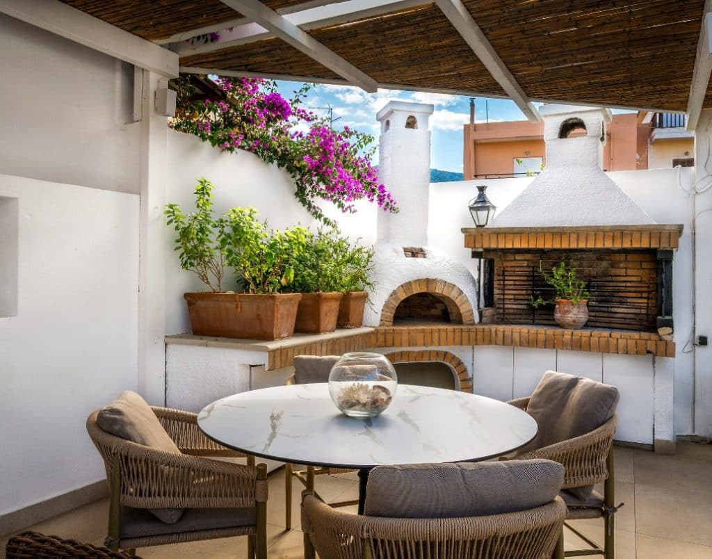 Outdoor entertainment area with a built-in brick oven, grill, and a dining table surrounded by cozy chairs under a pergola, adorned with vibrant bougainvillea and terracotta pots.