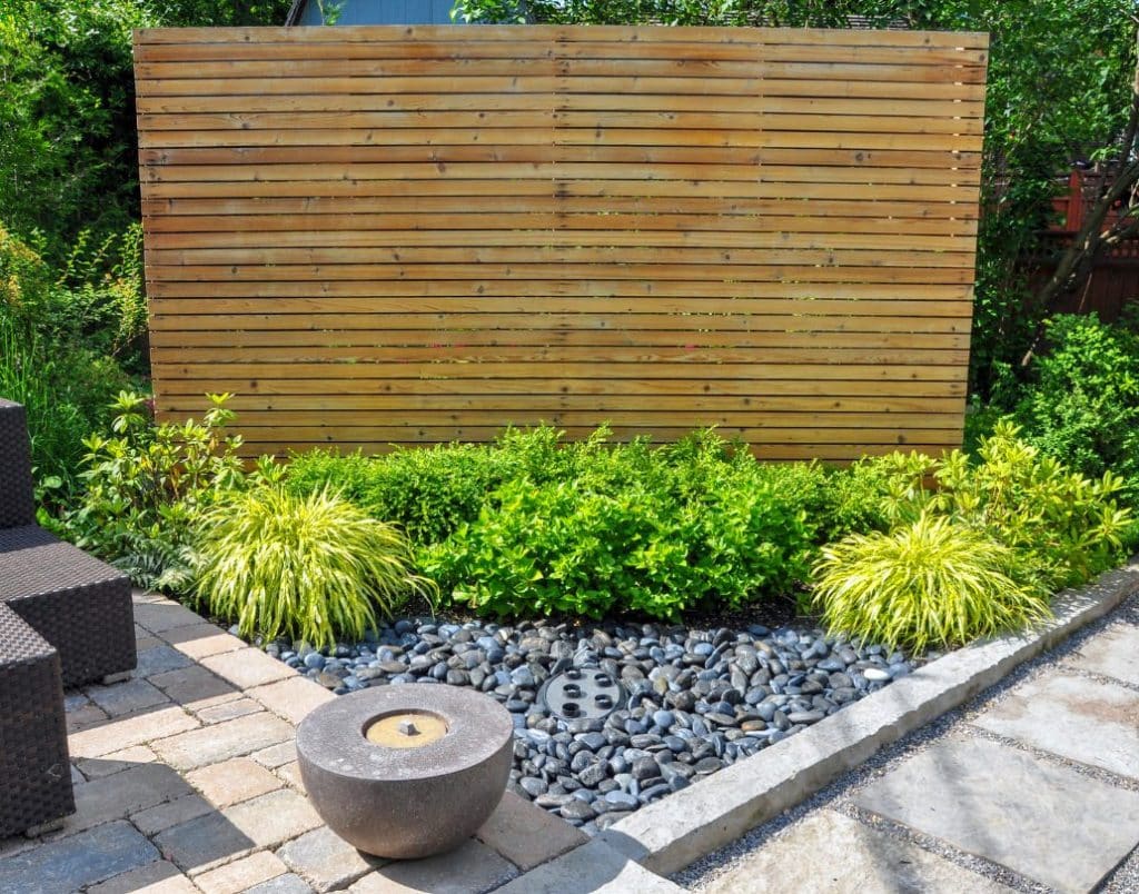 A wooden privacy screen in a landscaped backyard featuring ornamental plants and a modern water feature