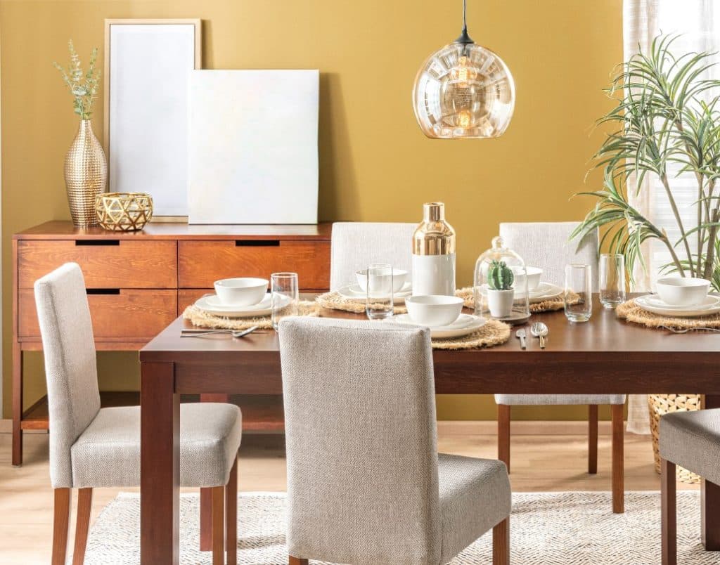 A dining room with a mustard yellow accent wall, a wooden table, and metallic decorative pieces.