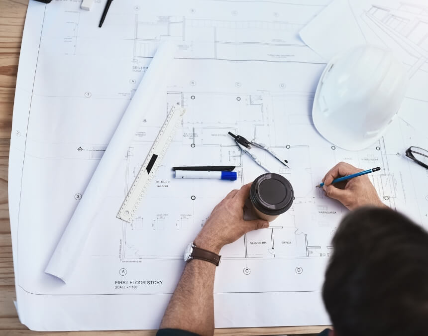 Person drafting architectural plans with a pen and coffee cup on a large blueprint spread across a desk.