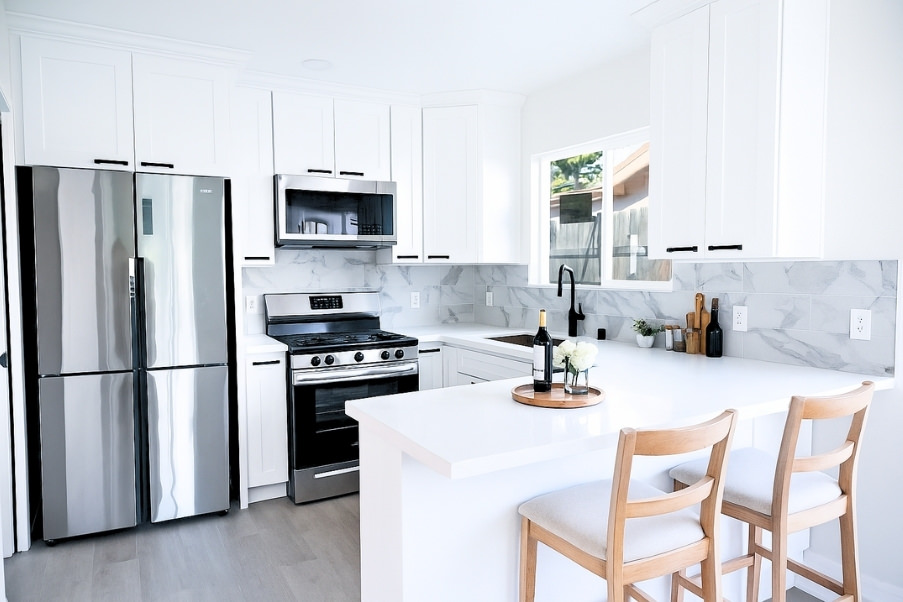 Bright coastal-inspired kitchen in garage conversion ADU with white cabinetry, marble backsplash, stainless steel appliances, and breakfast bar seating.