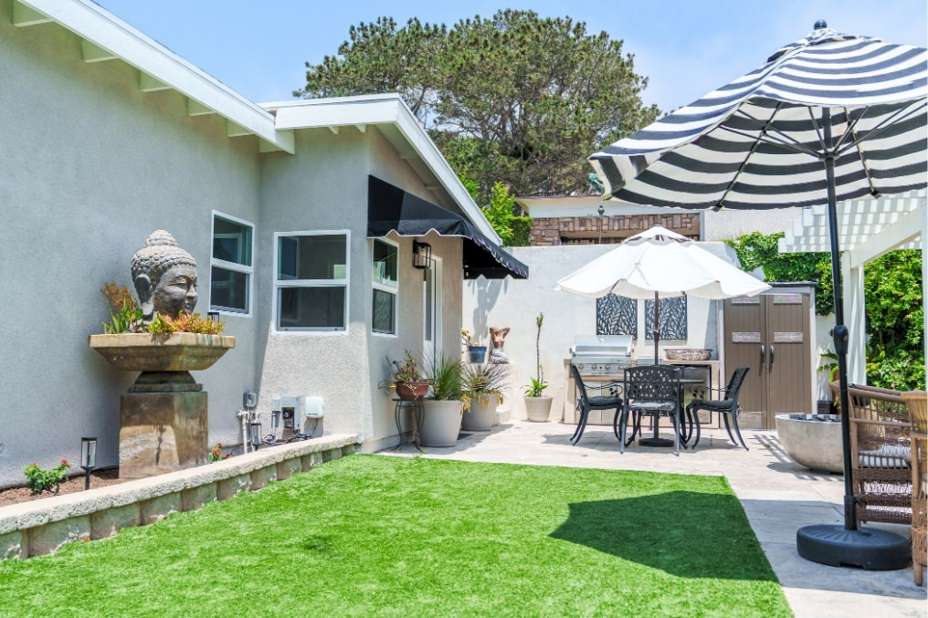 Tranquil boho-inspired backyard of detached ADU featuring lush turf, Buddha water fountain, outdoor dining area, and stylish umbrellas
