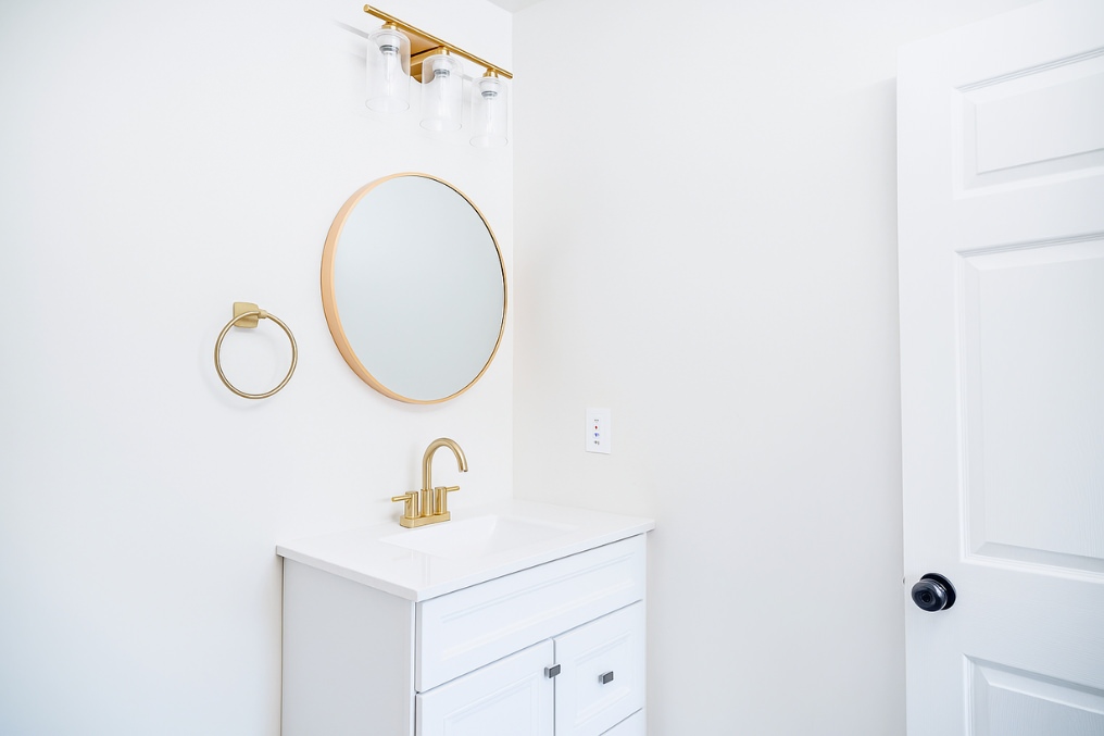 Coastal-inspired bathroom vanity with gold fixtures, round mirror, and minimalist decor in garage conversion.