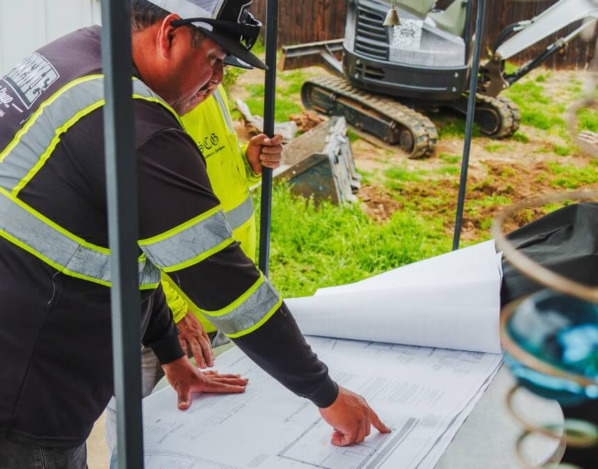 Contractors reviewing ADU plans on an active job site with equipment in the background