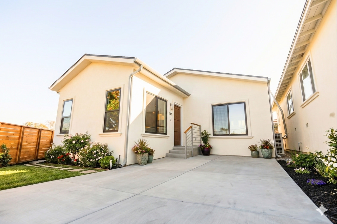 Front exterior of the Linda Vista Detached ADU featuring a modern cream stucco finish, large windows, a small entry staircase with railing, landscaped planters, and a spacious concrete driveway